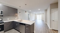 This contemporary single-floor residence showcases a thoughtfully designed open-concept layout that seamlessly connects the kitchen and living areas. The kitchen is a standout feature, equipped with dark wood-tone cabinetry, a light quartz countertop island with an undermount sink, subway tile backsplash, and a full suite of stainless steel appliances including a gas range, over-the-range microwave, dishwasher, and a French door refrigerator with an in-door ice dispenser. Stylish pendant lighting adds a modern accent above the island. The expansive living area is finished with light wood-look luxury vinyl plank flooring and neutral-toned walls, creating a bright and airy atmosphere. Recessed lighting throughout ensures consistent illumination, while a large sliding glass door invites natural light into the living space. The bedroom features plush wall-to-wall carpeting and a window with blinds for added comfort and privacy. The bathrooms are well-appointed — one features a dark-toned vanity with quartz countertop and modern fixtures, while the primary bath includes a large walk-in glass-enclosed shower with floor-to-ceiling tile work, built-in bench seating, and brushed gold hardware. A dedicated laundry area houses a full-size washer and dryer with overhead shelving, completing this well-equipped and modern living space.
