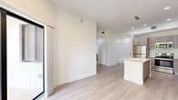 Located at Emerald Bay, this well-designed one-bedroom, one-bathroom unit offers a thoughtful layout across a single floor. Light wood flooring runs throughout, lending a clean, contemporary feel to every room.

The open-concept kitchen and living area forms the heart of the space. The kitchen is equipped with stainless steel appliances — including a refrigerator, range, and microwave — complemented by flat-front wood-toned cabinetry, white quartz countertops, and a central island with pendant lighting above. The kitchen flows seamlessly into the living area, which receives natural light through a sliding glass door that opens to an outdoor patio with lush greenery views.

The bedroom is a comfortable, well-proportioned room featuring a window with palm tree views, providing a serene atmosphere. It connects directly to a dedicated walk-in closet with wire shelving, offering generous storage. A hallway links the bedroom to the bathroom and the rest of the unit.

The bathroom is a standout feature, showcasing floor-to-ceiling marble-style tile in the glass-enclosed walk-in shower, a modern floating vanity with a white quartz countertop, and an illuminated LED mirror, creating a spa-like retreat. Recessed lighting throughout the unit adds to the bright, airy ambiance.