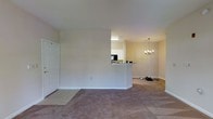 This single-floor residence offers a practical and welcoming open-concept layout designed for comfortable everyday living. Upon entry, a tiled foyer transitions into a spacious carpeted living area, providing a warm and inviting atmosphere throughout the main living space. A elegant chandelier hangs above the dining area, adding a refined touch to the open floor plan.

The kitchen features a peninsula-style counter with a pass-through design that connects seamlessly to the living and dining areas, promoting an open and social environment. White cabinetry lines the kitchen walls, complemented by overhead lighting that brightens the workspace. A black refrigerator is visible within the kitchen, reflecting a functional and well-equipped cooking space.

The interior is finished with neutral, warm-toned walls that enhance the sense of openness and allow natural light to reflect throughout the space. White trim and baseboards provide clean, classic detailing throughout. The combination of tile flooring at the entry and plush carpeting in the living areas balances practicality with comfort. Overall, the space presents a straightforward yet charming single-level layout well-suited for a variety of living arrangements.