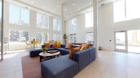 This contemporary communal space is defined by its striking double-height ceilings and an expansive, open floor plan that creates an immediate sense of grandeur upon entry. Large floor-to-ceiling windows and clerestory glazing flood the interior with natural light, while light-toned wood-look flooring runs throughout, lending warmth and continuity to the design.

The main lounge area centers around a generous modular navy sectional sofa accented with vibrant amber, rust, and yellow throw pillows, arranged around a round marble-top coffee table atop a rich red-toned area rug. A dramatic dark tile-clad fireplace with a linear flame serves as a bold focal point on one wall, grounding the space with texture and warmth.

Adjacent to the main lounge, a casual dining and social zone features sleek round dark-topped tables paired with modern yellow sculpted chairs on black wire frames, offering views of the surrounding exterior through large framed windows.

A dedicated lounge alcove showcases two pairs of mustard yellow high-back accent chairs flanking small cylindrical side tables, set against a gallery wall of large-scale abstract paintings in vivid, expressive color palettes—adding an artistic, curated character to the space.

The entry vestibule features glass double doors that open to the exterior, bathing the threshold in natural light. Throughout, the overall ambiance is bright, welcoming, and thoughtfully designed for social gathering.