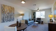 This single-floor space offers a thoughtfully designed open layout that seamlessly connects the dining and living areas, creating a cohesive and functional environment. The living area is carpeted and centers around a comfortable gray sofa paired with a tufted accent chair, complemented by a hairpin-leg coffee table that adds a contemporary touch. A ceiling fan overhead provides both comfort and airflow, while a large window invites generous natural light and offers views of the surrounding exterior. A tall decorative floor lamp and a lush potted plant add warmth and life to the space. The dining area flows naturally from the living room and features a round dark-wood table with white upholstered seating, anchored by an elegant overhead light fixture. The walls are painted in soft, neutral tones that enhance the bright and airy ambiance throughout. Decorative candles and abstract wall art contribute to a curated, welcoming atmosphere. The overall design balances modern simplicity with comfortable, livable details, making this a practical and inviting single-level space suited for everyday living and entertaining alike.