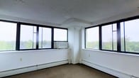 This single-floor space offers a bright and open layout defined by its standout corner window configuration. Large, black-framed windows wrap around two walls, flooding the interior with natural light and providing panoramic views of the lush, tree-lined surroundings outside — a refreshing visual connection to the outdoors. The elevated positioning of the unit enhances these views, giving the space an airy, elevated feel.

The interior features neutral white walls and a textured white ceiling, creating a clean and versatile backdrop suitable for a variety of uses. Carpeted flooring in a warm neutral tone adds a modest layer of comfort underfoot. Baseboard heating runs along the perimeter beneath the windows, providing practical climate control throughout the space.

Structural column detailing is visible in the corner area between the window banks, adding a subtle architectural element to the otherwise minimalist design. The overall ambiance is light-filled and functional, with the generous window presence being the defining characteristic of the space. Whether configured as an office, studio, or workspace, this unit offers a well-lit environment with pleasant outdoor views that contribute to a productive and comfortable atmosphere.