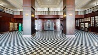 Located at 421 19th St in Moline, Illinois, this grand hallway is a striking example of classic early 20th-century civic architecture. The spacious, single-floor interior spans a generously proportioned footprint and immediately commands attention with its iconic black-and-white checkerboard terrazzo flooring, which extends throughout the entire space. Soaring double-height ceilings are adorned with ornate plasterwork and cornice detailing, while a wraparound mezzanine level with an elegant decorative railing overlooks the main hall below. Tall arched windows along the perimeter flood the space with abundant natural light, highlighting the richly crafted dark wood paneling that lines the walls and structural columns. A central entry vestibule features grand wood-framed glass doors and a built-in millwork surround, lending a formal, stately character to the entrance. Pendant light fixtures with green glass shades and wall-mounted brass sconces provide warm supplemental lighting throughout. Additional functional elements include a bank of individual mailboxes, elevator access, and a staircase leading to upper levels. The overall ambiance is one of timeless grandeur and architectural refinement, reflecting the building's distinguished heritage.