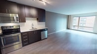 Located at 1111 East Union Street in Seattle's Capitol Hill neighborhood, this modern single-floor residence offers a thoughtfully designed open-concept layout that seamlessly connects the kitchen and living areas. The kitchen is outfitted with rich dark espresso cabinetry, sleek white countertops, stainless steel appliances including a refrigerator, electric range, built-in microwave, and dishwasher, all illuminated by track lighting overhead. Light wood-tone flooring flows throughout the main living area, complementing the neutral gray tones of the walls and creating a clean, contemporary aesthetic. A large window in the living area fills the space with natural light and provides views of the surrounding urban streetscape. The bedroom features soft carpeting, a generous closet with built-in shelving, and a calm, understated palette. The full bathroom is finished with tile flooring, a single-sink vanity with dark cabinetry and white countertop, and a full soaking tub with shower combination. The overall design emphasizes a modern, minimalist sensibility with consistent finishes throughout, making this a well-appointed urban residence in one of Seattle's most walkable and vibrant neighborhoods.