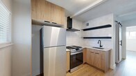 This thoughtfully designed 1-bedroom, 1-bathroom unit makes excellent use of its compact footprint, offering a cohesive blend of modern finishes and functional living spaces. Light hardwood flooring runs throughout, creating a warm, unified aesthetic from room to room.

The kitchen is a standout feature, equipped with stainless steel appliances including a gas range and full-size refrigerator. Warm wood-tone cabinetry — both upper and lower — is complemented by open black floating shelves and a tile backsplash, lending a contemporary yet inviting character. A sleek undermount sink with a matte black faucet adds a refined touch. A range hood overhead ensures practical ventilation.

The living and entry area is bright and open, with a window allowing natural light to filter in alongside the front door entry, keeping the space feeling airy despite its scale.

The bedroom is a serene, light-filled retreat with crisp white walls, a window with exterior views, and a closet that maximizes storage. Its clean, minimalist design offers a calm and restful atmosphere.

The bathroom elevates the unit with large-format marble-style wall tiles in the shower, dark floor tiles, a wall-mounted sink with gold fixtures, a round mirror, and a matte black shower fixture — all combining to create a spa-like aesthetic that punches well above its size.