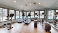 This fitness center is a bright and well-appointed space designed to support a comprehensive workout experience. The room features vaulted ceilings with recessed lighting, which contribute to an open and airy atmosphere. Large, multi-pane windows line the walls, flooding the interior with natural light and offering direct views of an outdoor swimming pool and surrounding grounds — an energizing backdrop for any workout session.

The flooring throughout is a warm-toned wood-look tile, adding a clean and contemporary feel to the space. The main exercise area is outfitted with a full suite of Precor cardio equipment, including multiple treadmills and elliptical cross-trainers, all positioned to take advantage of the pool views. A large wall-mounted mirror along one side of the room visually expands the space while allowing users to monitor their form.

The entry area features glass doors that open to the outdoors, providing easy access and excellent ventilation. Posted fitness center rules are clearly displayed near the entrance, and a stainless steel waste receptacle adds a polished, functional touch. The overall design is modern and well-maintained, creating an inviting environment that encourages regular use and supports an active lifestyle.