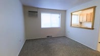 This single-story space presents a straightforward and functional interior. The main room visible features wall-to-wall carpet in a neutral brown-gray tone, providing a practical and comfortable underfoot surface. The walls are finished in a light beige, contributing to a clean, understated aesthetic throughout.

Natural light enters through a window fitted with horizontal blinds, offering both brightness and privacy control. A wall-mounted air conditioning unit is positioned above the window, serving as the primary cooling source for the room. A floor-level ventilation grille is also present, indicating a forced-air heating system beneath.

A pass-through window or open cutout with a wooden frame is visible on the adjacent wall, suggesting a connection to a neighboring room — likely a kitchen or secondary space — which adds a degree of openness to the layout.

The ceiling features a textured popcorn finish, a detail consistent with mid-century residential construction. Overall, the space is compact but adequately proportioned, with a neutral color palette and minimal architectural detailing that lends itself well to a variety of furnishing styles.
