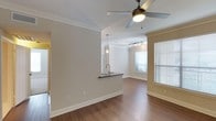 This single-floor residence offers a thoughtfully designed open-concept layout that promotes a sense of spaciousness and ease of movement throughout the living areas. Rich hardwood-style flooring runs continuously across the main living space, adding warmth and a cohesive aesthetic. Elegant crown molding frames the ceilings, lending a refined touch to the interior. The living area is bright and airy, benefiting from a large window dressed with white blinds that allows generous natural light to filter in. A ceiling fan with an integrated light fixture keeps the space comfortable and well-lit. Track lighting in the adjoining area provides additional illumination and a modern accent. The kitchen is connected to the main living area via a convenient pass-through counter with a dark contrasting surface, a stainless steel sink, and pendant lighting overhead — creating a functional and stylish point of interaction between the kitchen and living space. Neutral, warm-toned walls throughout complement the overall palette and provide a versatile backdrop. The layout flows naturally from one area to the next, making the space feel both functional and inviting for everyday living.