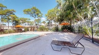 This outdoor pool area offers a classic Florida retreat, centered around a generously sized rectangular swimming pool with clear blue water and stainless steel entry rails. The surrounding deck is finished in light-toned tile, providing ample space for lounging and circulation. Multiple reclining strap lounge chairs are positioned throughout the deck, inviting relaxation in the open sunshine.

A wrought iron perimeter fence encloses the entire pool area, offering both security and visual openness to the verdant surroundings beyond. A life ring is mounted to the fence, reflecting attention to pool safety. An orange market umbrella with accompanying patio table and chairs is situated at one end of the deck, providing a shaded gathering spot.

The space is framed by mature tropical and native Florida vegetation, including tall slash pines, royal palms, and dense understory plantings that create a lush, natural backdrop. Low-rise residential buildings are visible in the background, set well beyond the fence line. The overall ambiance is relaxed and tropical, with abundant natural light and open sky views that are characteristic of a sunny Florida outdoor setting.