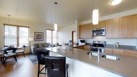 This thoughtfully designed single-floor space showcases an open-concept layout that effortlessly connects the kitchen, dining, and living areas into one cohesive and functional environment. The kitchen is a standout feature, outfitted with warm honey-toned cabinetry, stainless steel appliances including an electric range and built-in microwave, and a sleek countertop that extends into a generous breakfast bar. Two stylish leather bar stools invite casual dining and conversation, while elegant cylindrical pendant lights hang overhead, casting a warm and inviting glow. The countertops carry a neutral, stone-like finish that complements the overall color palette of the space. The open living area flows naturally from the kitchen, offering room for comfortable seating and accented by natural light streaming through horizontal blinds. Light hardwood-style flooring runs throughout, enhancing the sense of continuity and warmth. Tasteful décor, including framed artwork and soft textiles, adds character without overwhelming the clean, modern aesthetic. The high ceilings amplify the feeling of openness, making the space feel airy and expansive. A hallway visible in the background suggests additional rooms, completing this well-balanced and welcoming home.