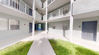 This ground-floor unit is part of a well-maintained multi-story residential building with a distinctive open-air courtyard design. The exterior common areas are defined by clean concrete walkways flanked by neatly trimmed grass strips, creating an inviting approach to each unit's entrance. The building's stucco facade is finished in a light grey-green tone, complemented by crisp white railings that line the covered balconies on the upper levels, giving the structure a clean and cohesive aesthetic. Each unit on the ground floor features a solid entry door in a deep charcoal hue, providing a strong visual contrast against the light exterior walls. Overhead, the covered corridor ceiling offers shade and shelter along the walkway, while structural columns define the open-air passageway that connects through to the building's interior courtyard. The upper floors feature wrap-around balconies with white metal railings, adding a layered, open feel to the building's architecture. The ground-floor interior spaces, while limited in captured detail due to lighting conditions, appear to include a bathroom and additional rooms typical of a residential unit. The overall ambiance of the property is clean, functional, and well-kept, characteristic of a classic Florida-style multi-unit residential building.