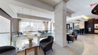 This refined communal space exudes a contemporary elegance defined by clean lines, premium materials, and a carefully curated aesthetic. Towering marble-clad columns serve as dramatic architectural focal points, lending a sense of grandeur to the open-plan layout. The soaring ceilings amplify the sense of volume and airiness throughout the space.

Floor-to-ceiling windows flood the interior with an abundance of natural light, framing views of mature trees and the surrounding landscape, creating a seamless connection between the interior and the natural environment outside. Sheer drapery softens the light while maintaining the bright, welcoming atmosphere.

Stylish globe pendant lights hang in clusters, adding warmth and a sculptural quality to the lounge area. Dark upholstered armchairs and accent tables are thoughtfully arranged to encourage conversation and relaxation, while the neutral toned carpet grounds the space with a quiet sophistication.

A glass partition defines a secondary seating and meeting area visible beyond, offering both openness and subtle separation of function. The overall ambiance is one of polished refinement — a professional yet comfortable environment that balances modern design sensibility with an inviting, hospitality-inspired warmth.