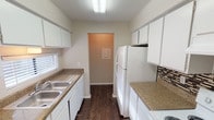 Located at 7500 Wicker Dr in Fort Worth, TX, this single-story unit offers a functional and straightforward layout suited for comfortable everyday living. The galley-style kitchen is equipped with white cabinetry providing ample storage, granite-look countertops, a double stainless steel sink, and a white refrigerator. Dark hardwood-style flooring runs throughout the kitchen, complemented by a mosaic tile backsplash accent and a window that allows natural light to filter in. The kitchen connects directly to an adjacent utility or storage area.

The living area features the same dark hardwood-style flooring, a textured ceiling, and a large sliding glass door fitted with vertical blinds that floods the space with natural light. The bedrooms are carpeted and include ceiling fans with light fixtures, windows with blinds for natural light, and closets offering practical storage. Neutral wall tones throughout create a clean, understated aesthetic.

The bathroom includes a tub and shower combination with tiled surround, dark hardwood-style flooring, a vanity with granite-look countertop and integrated sink, white cabinetry above, and a Hollywood-style light bar above the mirror. Overall, the unit presents a tidy, move-in-ready interior with consistent finishes throughout.