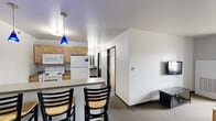 Located at 1284 Midway Rd in Menasha, WI, this single-floor unit offers four bedrooms and two bathrooms within a practical, efficiently organized layout. The combined kitchen and living area serves as the heart of the space, featuring warm honey-toned cabinetry, a granite-style countertop peninsula with bar seating for three, and eye-catching blue pendant lights overhead. White appliances — including a refrigerator, electric range, microwave, and dishwasher — complete the kitchen. The adjacent living area provides a comfortable lounge space with a wall-mounted TV and a window that brings in natural light. Each of the four bedrooms is carpeted and simply appointed, with individual windows offering views of the surrounding greenery. The bedrooms are compact yet functional, each capable of accommodating a bed and small furnishings. The two bathrooms differ in style: one features a floating vanity with a round sink and mirror, while the other is a more spacious, accessibility-friendly room with grab bars and vinyl tile flooring. A hallway and closet round out the floorplan, contributing to the unit's straightforward and utilitarian design throughout.