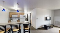 Located at 1284 Midway Rd in Menasha, WI, this single-floor unit offers four bedrooms and two bathrooms within a practical, efficiently organized layout. The combined kitchen and living area serves as the heart of the space, featuring warm honey-toned cabinetry, a granite-style countertop peninsula with bar seating for three, and eye-catching blue pendant lights overhead. White appliances — including a refrigerator, electric range, microwave, and dishwasher — complete the kitchen. The adjacent living area provides a comfortable lounge space with a wall-mounted TV and a window that brings in natural light. Each of the four bedrooms is carpeted and simply appointed, with individual windows offering views of the surrounding greenery. The bedrooms are compact yet functional, each capable of accommodating a bed and small furnishings. The two bathrooms differ in style: one features a floating vanity with a round sink and mirror, while the other is a more spacious, accessibility-friendly room with grab bars and vinyl tile flooring. A hallway and closet round out the floorplan, contributing to the unit's straightforward and utilitarian design throughout.