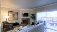 This single-floor residence offers a thoughtfully designed living space that balances comfort and contemporary style. The living room serves as the heart of the home, anchored by a classic stone fireplace with a clean white mantle and a wall-mounted flat-screen TV above, creating a natural focal point for the room. Crown molding lines the ceiling, adding an elegant architectural detail throughout. Large-scale abstract artwork adorns the walls, contributing to the space's curated aesthetic. Natural light pours in generously through a full-length sliding glass door fitted with vertical blinds, offering views of and access to an outdoor patio or balcony area. The room is finished with warm wood-tone flooring, complemented by a layered area rug with a bold geometric pattern that grounds the seating arrangement. A plush, neutral-toned sofa and coordinating accent seating fill the space comfortably, while a round white coffee table adds a modern touch. A tall indoor tree plant introduces a refreshing natural element. The overall ambiance is clean, modern, and inviting, with a neutral color palette of warm whites and soft grays that lends the space a timeless and versatile character.