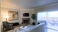 This single-floor residence offers a thoughtfully designed living space that balances comfort and contemporary style. The living room serves as the heart of the home, anchored by a classic stone fireplace with a clean white mantle and a wall-mounted flat-screen TV above, creating a natural focal point for the room. Crown molding lines the ceiling, adding an elegant architectural detail throughout. Large-scale abstract artwork adorns the walls, contributing to the space's curated aesthetic. Natural light pours in generously through a full-length sliding glass door fitted with vertical blinds, offering views of and access to an outdoor patio or balcony area. The room is finished with warm wood-tone flooring, complemented by a layered area rug with a bold geometric pattern that grounds the seating arrangement. A plush, neutral-toned sofa and coordinating accent seating fill the space comfortably, while a round white coffee table adds a modern touch. A tall indoor tree plant introduces a refreshing natural element. The overall ambiance is clean, modern, and inviting, with a neutral color palette of warm whites and soft grays that lends the space a timeless and versatile character.