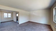 Located at 835 Blooming Grove Turnpike in New Windsor, New York, this single-floor space offers a clean and straightforward layout designed for practical use. The main living area is generously proportioned, featuring wall-to-wall gray carpeting that provides a neutral, comfortable foundation throughout. Smooth white walls and a plain white ceiling create a bright, open atmosphere, while a baseboard heating unit along the perimeter ensures efficient climate control. Natural light filters in through a window visible in the adjacent room, contributing to an airy feel. A wide rectangular doorway connects the main living area to a secondary room, which also features carpeted flooring and window access, allowing for flexible use as an additional living space, office, or sleeping area. The overall design is minimal and move-in ready, offering a blank canvas that can be easily personalized. The clean lines, neutral color palette, and open flow between rooms make this a versatile and functional space suited to a variety of needs.