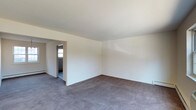 Located at 835 Blooming Grove Turnpike in New Windsor, New York, this single-floor space offers a clean and straightforward layout designed for practical use. The main living area is generously proportioned, featuring wall-to-wall gray carpeting that provides a neutral, comfortable foundation throughout. Smooth white walls and a plain white ceiling create a bright, open atmosphere, while a baseboard heating unit along the perimeter ensures efficient climate control. Natural light filters in through a window visible in the adjacent room, contributing to an airy feel. A wide rectangular doorway connects the main living area to a secondary room, which also features carpeted flooring and window access, allowing for flexible use as an additional living space, office, or sleeping area. The overall design is minimal and move-in ready, offering a blank canvas that can be easily personalized. The clean lines, neutral color palette, and open flow between rooms make this a versatile and functional space suited to a variety of needs.