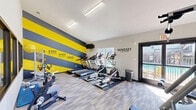 This dedicated fitness center at 5151 Highland Rd in Baton Rouge, Louisiana, is a well-equipped, single-room exercise facility designed to support a comprehensive workout routine. The space features elevated ceilings that contribute to an open, airy atmosphere, while large windows along multiple walls invite abundant natural light and offer views of the surrounding outdoor area, including a pool visible from within the room.

The flooring consists of stylish patterned carpet tiles in gray and yellow tones, providing both comfort and durability underfoot. Cardio equipment is prominently featured, including treadmills, an elliptical trainer, and stationary cycling bikes, all positioned to take advantage of the natural light and views. A wall-mounted television is conveniently placed for entertainment during workouts.

Strength training is well accommodated with a multi-station cable machine and a variety of bench press and resistance equipment. Full-length mirrors on one wall enhance the sense of space and allow users to monitor their form. A bold accent wall decorated with bold yellow and dark navy diagonal stripes and motivational phrases such as "Stay Positive," "Work Hard," and "Mindset Is Everything" energizes the environment. Double glass doors provide easy access and connect the room to the exterior. Overall, the space offers a focused, motivating environment for fitness.