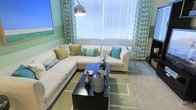 This single-floor living space is designed around a cohesive green and white color palette that creates a fresh, inviting atmosphere. The living area serves as the focal point, anchored by a large cream-colored sectional sofa adorned with an assortment of decorative pillows in shades of green, teal, and white, adding layers of texture and visual interest. A bold arc floor lamp arches over the seating area, providing both functional and ambient lighting.

The room benefits from a generous window dressed with white horizontal blinds and framed by green floral patterned curtains, allowing ample natural light to fill the space. A dark-finished rectangular coffee table sits centrally on a geometric green and beige area rug, styled with decorative vases and accents that complement the room's color story.

The walls feature a two-tone green horizontal stripe design, adding depth and character to the space. A framed piece of artwork adorns one wall, reinforcing the room's nature-inspired aesthetic. The overall ambiance is relaxed yet well-curated, blending comfort with thoughtful decorative detail.