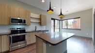 Located at 1538 Eighth Ave in Greeley, Colorado, this single-floor unit delivers a well-appointed, modern living environment with thoughtfully selected finishes throughout. The open-concept kitchen serves as a central gathering point, featuring light wood cabinetry, dark countertops, a classic white subway tile backsplash, and a full suite of stainless steel appliances including a refrigerator, range, microwave, and dishwasher. A central island with a contrasting white base and gray countertop provides additional prep space and casual seating. Stylish pendant lighting in a dark bronze finish anchors the kitchen visually, while large windows allow natural light to flow in and offer views of the surrounding streetscape.

Warm hardwood-style flooring extends consistently throughout the unit, contributing to a cohesive and contemporary aesthetic. The bedrooms are bright and well-proportioned; one features a sliding barn-style door that opens to a walk-in closet with built-in shelving. Two bathrooms serve the unit—one includes a soaking tub with full white subway tile surround and dark hardware, while the other features a light wood vanity with dark countertop, framed mirror, and tiled shower. In-unit laundry connections are also present, adding to the practical functionality of this well-designed space.