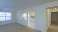 This single-floor space offers a clean, minimalist layout designed for comfortable everyday living. The main living area features neutral carpet flooring and smooth white walls that reflect natural light, creating a bright and airy atmosphere. A pass-through window connects the living area to the adjacent kitchen, enhancing the sense of openness between the two spaces. The kitchen is accessible via a wide doorway and is finished with light tile flooring, white cabinetry, dark countertops, and a built-in dishwasher. Natural light filters into the kitchen through vertical-blind-covered windows, adding warmth to the space. The white ceiling throughout maintains a consistent, clean aesthetic. The overall design is straightforward and functional, with a neutral color palette that offers a versatile backdrop for a variety of interior styles. The seamless flow between the living and kitchen areas makes the space feel connected and practical for daily use.