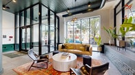 Located at 780 S 52nd St in the Cobbs Creek neighborhood of Philadelphia, this expansive ground-floor lobby blends industrial and contemporary design to create a striking first impression. The space is anchored by a prominent structural column with a dark metal finish, which complements the exposed ductwork on the painted ceiling above. Large, floor-to-ceiling steel-framed windows flood the interior with abundant natural light and offer open views of the surrounding streetscape and tree canopy.

The entry corridor features large-format tile flooring and is accented by a bold neon "1" wall installation, establishing a distinctive sense of identity upon arrival. A set of wide, low-profile stairs with sleek metal railings leads up to a hallway finished with deep forest-green paneled walls and warm wall sconces, where two elevator bays provide vertical access throughout the building.

A carefully curated lounge area is defined by a rich, patterned area rug, a caramel leather sofa, sculptural pendant lighting, and a mix of modern accent chairs arranged around a cylindrical stone coffee table. Industrial-style steel shelving units and lush tropical plants add warmth and texture. Steel-framed glass partitions elegantly divide the lounge from the circulation paths, maintaining visual openness throughout this thoughtfully designed communal space.