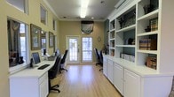 This single-floor business center offers a thoughtfully designed workspace that balances functionality with a welcoming aesthetic. The room is finished with warm yellow-green walls accented by crisp white crown molding, creating a clean and professional atmosphere. Light hardwood flooring runs throughout, adding warmth and a sense of openness to the space.

Workstations line both sides of the room, each equipped with computer monitors and ergonomic task chairs, providing ample workspace for multiple users simultaneously. A glass-paneled exterior door at the far end of the room invites natural light deep into the space, complemented by overhead fluorescent lighting to ensure consistent illumination throughout the day.

On one side, an expansive built-in shelving and cabinet unit in white offers generous storage and display space, housing books, decorative objects, and organizational materials. The cabinetry below provides additional closed storage, keeping the space tidy and uncluttered.

Decorative touches — including ornate mirrors, framed artwork, and carefully placed accessories — lend a refined character to what might otherwise be a purely utilitarian room. The overall ambiance is one of quiet efficiency, making this business center a practical and pleasant environment for work and research.