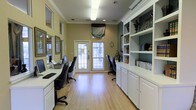 This single-floor business center offers a thoughtfully designed workspace that balances functionality with a welcoming aesthetic. The room is finished with warm yellow-green walls accented by crisp white crown molding, creating a clean and professional atmosphere. Light hardwood flooring runs throughout, adding warmth and a sense of openness to the space.

Workstations line both sides of the room, each equipped with computer monitors and ergonomic task chairs, providing ample workspace for multiple users simultaneously. A glass-paneled exterior door at the far end of the room invites natural light deep into the space, complemented by overhead fluorescent lighting to ensure consistent illumination throughout the day.

On one side, an expansive built-in shelving and cabinet unit in white offers generous storage and display space, housing books, decorative objects, and organizational materials. The cabinetry below provides additional closed storage, keeping the space tidy and uncluttered.

Decorative touches — including ornate mirrors, framed artwork, and carefully placed accessories — lend a refined character to what might otherwise be a purely utilitarian room. The overall ambiance is one of quiet efficiency, making this business center a practical and pleasant environment for work and research.