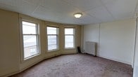 This single-floor space presents a straightforward yet characterful interior with several notable architectural and functional details. The main room is anchored by a distinctive bay window configuration, featuring three double-hung windows arranged in an angled formation that curves outward, allowing natural light to fill the room from multiple angles while offering views of the surrounding urban streetscape. The carpeted flooring throughout adds a layer of warmth and comfort to the room, and the neutral, cream-toned walls provide a clean, versatile backdrop. A drop tile ceiling with a centrally mounted flush light fixture provides even overhead illumination. A classic cast-iron radiator positioned along the wall is a standout period detail, indicating a traditional hot-water or steam heating system characteristic of older construction. The space has a simple, uncluttered layout with functional proportions, offering flexibility for various configurations. Overall, the space combines historic architectural elements, including the bay windows and radiator, with a clean, open interior that would suit a range of uses.