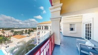 Located at 401 Northeast Mizner Boulevard in the heart of Boca Raton, Florida, this outdoor balcony space offers a compelling vantage point above the city's distinctive Mediterranean Revival streetscape. The balcony is defined by its ornate architectural detailing, including decorative columns with classic molding and cream-colored stucco finishes that reflect the building's elegant design heritage. A bold red and black metal railing frames the perimeter, providing both safety and a striking visual contrast against the balcony's smooth blue-painted concrete floor. The space is compact yet purposeful, comfortably accommodating outdoor seating — as evidenced by the presence of a modern chair — making it an ideal spot for relaxing and taking in the surroundings. The elevated position affords open views of Boca Raton's urban landscape, with neighboring Mediterranean-style buildings, terracotta rooftops, swaying palm trees, and lush greenery stretching into the distance beneath wide, sun-drenched Florida skies. Abundant natural light bathes the balcony throughout the day, enhancing the warm, resort-like ambiance that is characteristic of this premier Boca Raton address. This outdoor space serves as a refined extension of the building's architectural identity.