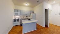 Located at 4395 Birchwood Dr in Wilmington, NC, this single-floor residence offers a thoughtfully laid-out living space with a clean, neutral aesthetic throughout. The kitchen is a functional highlight of the home, featuring gray cabinetry, white appliances including an electric range, built-in microwave, refrigerator, and dishwasher, along with a double stainless steel sink and light countertops. A peninsula breakfast bar extends the prep and dining space, making it ideal for casual meals or entertaining. Warm hardwood-style flooring runs through the kitchen and common areas, adding a welcoming feel to the space. The open layout, visible in the aerial dollhouse view, connects the kitchen seamlessly to the main living area, which features plush carpeting underfoot. The bedroom is generously sized with soft carpet flooring and a wide set of windows that frame lush greenery outside, flooding the room with natural light. The bathroom is neatly appointed with a gray vanity, large mirror, and hardwood-style flooring, providing a cohesive design language that ties the home together. Bright white walls and clean lines throughout give the space a fresh, move-in-ready feel.