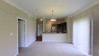 This single-floor space showcases an open-concept design that seamlessly connects the living area and kitchen. The living space is finished with plush, light-toned carpet that extends throughout, complemented by neutral beige walls and white crown molding that add a clean, polished look to the interior.

The kitchen serves as a functional centerpiece, featuring dark espresso cabinetry that contrasts with light stone countertops along the breakfast bar peninsula. A decorative tile backsplash adds texture behind the sink area, while stainless steel appliances, including a refrigerator, contribute a modern touch. A stylish pendant light hangs above the breakfast bar, providing focused task lighting, while a flush-mount ceiling fixture illuminates the adjoining area.

A structural column near the kitchen entry adds an architectural element to the space. A hallway visible beyond the kitchen leads to additional rooms, suggesting a thoughtfully segmented floor plan. Doorways on either side of the living area provide access to other parts of the space, enhancing the overall flow and functionality of the layout.