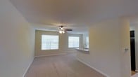 This single-floor residence offers a well-designed, open-concept layout that seamlessly connects the living area to the kitchen. The living space is finished with soft, plush wall-to-wall carpeting and features a ceiling fan with integrated lighting, providing both comfort and air circulation. Recessed lighting in the ceiling adds supplementary illumination throughout the space.

Natural light is a defining characteristic of this home, with multiple windows fitted with white horizontal blinds allowing for adjustable light control. The windows face outward to a lush green exterior, bringing a sense of the outdoors inside.

The kitchen is integrated into the open floor plan via a peninsula-style counter that doubles as a divider between the living and cooking areas. A single-basin sink is positioned at the peninsula, making it a practical and social focal point. The neutral color palette — from the beige walls and white trim to the light-toned carpet — gives the entire space a clean, cohesive, and inviting aesthetic. The consistent use of neutral tones throughout provides a versatile backdrop suitable for a wide range of interior design preferences.