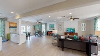 This single-floor space functions as a welcoming community or leasing center, thoughtfully designed with an open floor plan that encourages flow between distinct functional zones. Light wood-style flooring spans the entire area, lending a clean, cohesive foundation that complements the neutral gray and beige wall tones throughout.

The main lounge area serves as the centerpiece, furnished with a gray sectional sofa, a bold teal accent chair, and a dark wood coffee table anchored by a vibrant orange and teal geometric area rug. A chevron-patterned shiplap accent wall frames a wall-mounted flat-screen television and a walnut media console, adding a contemporary architectural detail to the space.

Adjacent to the lounge, a dining area features both a round table with woven chairs and a counter-height bar table with green seating, providing flexible gathering options. French doors in this area open directly to an outdoor space, allowing ample natural light to filter in alongside the multiple large windows dressed in teal patterned curtains.

Ceiling fans installed throughout ensure comfortable airflow across the open layout. Recessed lighting and decorative table lamps complement the natural light. Colorful artwork and decorative accessories in teal, orange, and amber tones give the space a lively, resort-inspired ambiance.