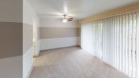 This single-floor unit offers a well-organized layout with multiple rooms designed for comfortable everyday living. The main living area is generously proportioned, featuring soft carpeted flooring, a textured ceiling, and a ceiling fan with a built-in light fixture. A set of large vertical-blind-covered sliding glass doors floods the space with natural light and provides access to an outdoor area. The accent wall, painted in two-toned tan and beige horizontal bands, adds a subtle design detail to the room.

The bedrooms are carpeted and feature textured ceilings, neutral white walls, and windows with vertical blinds that let in natural light and offer outdoor views. One of the bedrooms benefits from direct access to both a walk-in closet—complete with a clothing rod and ample storage—and a full bathroom, making it a practical private retreat.

The bathroom is clean and efficiently arranged, featuring a glass-door walk-in shower, a toilet, and a single-sink vanity topped with chrome fixtures. A dark-framed wall mirror and a matching dark-wood wall cabinet with open shelving provide functional storage while adding a contrasting design element to the otherwise light and bright space. Mosaic tile flooring in the bathroom area completes the look with a classic touch.