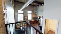 This Germantown Mill Loft showcases a striking blend of industrial heritage and contemporary living. The space is defined by its dramatic ceiling height, where original wooden ceiling planks and large exposed HVAC ductwork serve as standout architectural features that pay homage to the building's mill origins. Weathered exposed brick walls run throughout, adding texture and character to the interior.

The open-concept layout allows the kitchen and living areas to flow seamlessly into one another. The kitchen is equipped with stainless steel appliances, including a range and refrigerator, complemented by warm wood cabinetry and a sink with countertop workspace. Pendant lighting hangs above, providing functional illumination. The living area features a comfortable seating arrangement, with natural light streaming in through multiple large windows fitted with blinds, offering views to the outside while brightening the entire space.

Hardwood-style flooring runs throughout the main level, adding warmth against the industrial backdrop. An interior railing overlooks the open floor plan below, emphasizing the loft's generous vertical scale and creating a sense of openness throughout.