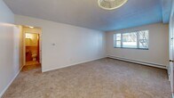 Located at 209 Pleasant Street in Melrose, MA, this one-bedroom, one-bathroom unit offers a practical and well-organized single-floor layout. The combined living and dining area is generously proportioned, featuring wall-to-wall carpeting, neutral white walls, and a warm wood-toned entry door that anchors the space. A pendant light fixture and ceiling fan provide overhead lighting, while baseboard heating units run along the exterior walls. A window in the living area invites natural light and outdoor views, enhancing the open feel of the room. The kitchen is efficiently designed with white shaker-style cabinetry, light countertops with wood-toned edging, a stainless steel sink, and a white gas range — all set on light hardwood flooring that adds warmth to the space. A carpeted hallway connects the main living area to the bedroom and bathroom, with multiple closets providing ample storage throughout. The spacious bedroom accommodates a comfortable furniture arrangement and includes a set of built-in bifold closet doors, carpeted flooring, and a window that fills the room with natural light. The bathroom features classic pink tile accents. Overall, the unit offers a clean, functional layout suited for everyday living.