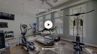 Located at 28155 Encanto Drive in Menifee, CA, this fitness center offers a well-appointed exercise space designed to support a comprehensive workout routine. The room is bathed in natural light streaming through large, white-framed windows that line multiple walls, offering pleasant views of the surrounding pool area and palm tree-dotted landscape.

The interior is finished with soft sage-green walls and a distinctive checkered carpet tile floor in neutral tones, creating a clean and energizing atmosphere. Multiple ceiling fans are mounted throughout, ensuring consistent airflow and comfort during workouts.

The space is outfitted with a robust selection of equipment, including treadmills, elliptical trainers, a recumbent bike, and a cable/weight machine for strength training. A dumbbell rack with a full range of free weights, wall-mounted exercise mats, and stability balls of various sizes round out the functional offerings. A wall-mounted flat-screen television provides entertainment during cardio sessions.

A full-length mirror along one wall enhances the sense of space while allowing users to monitor their form. An adjacent room, visible through an open doorway, appears to serve as a supplementary area. The overall ambiance is bright, open, and purposefully designed for active use.