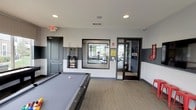 This single-floor amenity space is designed to support both recreation and fitness, offering residents a well-equipped environment for leisure and activity. The billiards room serves as the centerpiece, featuring a full-size pool table with a grey felt surface, complemented by a wall-mounted cue rack stocked with billiard sticks and a full set of balls. The walls are finished in a two-tone design, with light grey upper sections and a darker grey lower band, creating a modern and cohesive aesthetic. Recessed ceiling lighting provides even, functional illumination throughout the space. Carpeted flooring in a neutral tone adds comfort underfoot. A large interior window connects the billiards room to the adjacent fitness center, offering a clear view into the workout area, which is accessible through glass-paneled doors. The fitness center is visible beyond, outfitted with exercise equipment and bathed in natural light from exterior windows. Signage and directional elements are thoughtfully placed throughout, contributing to the organized and purposeful layout of the space. Overall, the amenity area presents a clean, contemporary design that balances active recreation with community-oriented living.