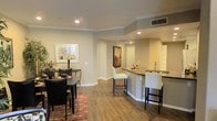 This single-floor residence showcases an open-concept layout that integrates the dining area and kitchen into one cohesive living space. The flooring throughout is a warm hardwood-style material that adds continuity and a polished feel to the interior.

The dining area is positioned near the entry of the space, accommodating a round table with dark chairs and a patterned area rug that anchors the zone. Crown molding runs along the ceiling perimeter, adding a refined architectural detail to the room.

The kitchen features a curved breakfast bar with dark countertops and seating for two high-back bar stools, making it functional for both casual dining and food preparation. A sink is integrated into the bar counter, and recessed lighting above ensures the workspace is well-lit. The kitchen opens further toward what appears to be an additional utility or laundry area visible in the background.

Neutral-toned walls, white baseboards, and ceiling vents are consistent throughout, contributing to a clean, well-maintained aesthetic. The open sightlines between rooms enhance the sense of space, making the interior feel airy and connected.