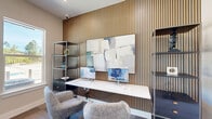 Located at 2300 N Loop 336 W in Conroe, Texas, this thoughtfully designed single-room workspace delivers a polished and functional environment within a compact footprint. The room's most defining architectural feature is a full-height slatted wood accent wall, which adds warmth, texture, and a contemporary aesthetic to the space. Against this backdrop, a sleek floating white desk anchors the work area, accompanied by stylish upholstered swivel chairs that blend comfort with modern design. Flanking the desk are open metal shelving units with built-in drawer storage, providing practical organization without sacrificing visual appeal. Abstract wall art complements the neutral color palette, adding a creative touch to the professional setting. The flooring features a patterned carpet tile that introduces subtle variation underfoot, while clean white walls and white trim maintain a bright, airy atmosphere throughout. Two windows on the far wall invite natural light and offer views of the surrounding outdoor landscape, including greenery and open sky. A frosted-glass interior door and louvered closet doors add refined architectural detail to the entry area. Recessed lighting ensures consistent illumination across the space, making it well-suited for productivity throughout the day.
