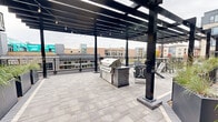 Located in the heart of downtown Eau Claire, Wisconsin, this contemporary outdoor terrace offers a well-appointed communal gathering space designed for both relaxation and entertainment. The paver-tiled deck is anchored by a striking black steel pergola fitted with overhead string lights, creating an inviting ambiance day and night. A built-in masonry grill station serves as the centerpiece of the outdoor kitchen area, complemented by Adirondack-style seating and high-top bar tables positioned throughout the space. A dedicated lounge section features cushioned chaise lounges set atop a lush artificial turf area, providing a resort-like retreat. Large, modern planter boxes filled with ornamental grasses add a natural, sculptural element and define distinct zones within the terrace. Black metal railing borders the perimeter, offering unobstructed views of the surrounding Eau Claire cityscape. The exterior building facade features contemporary horizontal siding in neutral tones, with large glass doors and windows that connect the indoor and outdoor environments seamlessly. The overall design blends urban sophistication with comfortable, functional outdoor living.