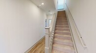 This well-appointed three-story home in Huntsville, Alabama offers four bedrooms, five bathrooms, and a thoughtfully designed layout spread across three distinct floors. The ground level houses an attached garage with a contemporary glass-panel door, an electrical panel, and direct interior access via carpeted staircases with clean white railings and wood-look flooring in the stairwells. The main living floor serves as the heart of the home, featuring an open-concept kitchen and living area. The kitchen is equipped with two-tone wood cabinetry, a large center island with a dark stone countertop, stainless steel appliances, and modern pendant lighting — all anchored by warm hardwood-style flooring. The upper floor hosts the bedrooms, including a generously sized primary suite with plush carpeting, a ceiling fan, a wide window offering outdoor views, and a walk-in closet with wire shelving. Bathrooms throughout feature large-format tile surrounds, sleek matte black hardware, wood-toned vanities, and granite countertops. A dedicated laundry room on the upper level includes side-by-side washer and dryer units, a utility shelf, and a bright window. The home's clean, modern aesthetic is consistent throughout, with white walls, recessed lighting, and durable flooring choices that balance style and practicality.
