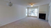 This single-floor space offers a clean, neutral aesthetic designed for comfortable everyday living. The main living area is generously sized, featuring soft wall-to-wall carpeting that adds warmth underfoot. Large windows fitted with horizontal blinds allow abundant natural light to fill the room, offering views of the surrounding greenery outside. A ceiling fan with integrated lighting is centrally positioned, providing both airflow and illumination to the space. A fireplace anchors one side of the room, serving as a natural focal point that adds character and a cozy ambiance. The walls are finished in a soft, neutral tone that complements the light carpeting, creating a cohesive and inviting atmosphere throughout. The open layout of the living area allows for flexible furniture arrangements and provides a sense of spaciousness. Overall, the space presents a straightforward, functional design with key comfort features — natural light, a fireplace, and practical finishes — making it a welcoming and versatile environment.