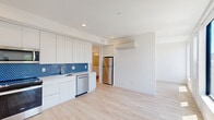 This thoughtfully designed studio presents a bright, contemporary living space spread across a single floor. The open-plan kitchen and living area is anchored by a sleek, modern kitchen outfitted with floor-to-ceiling white cabinetry, a bold cobalt blue penny-tile backsplash, and a full suite of stainless steel appliances including a range, over-the-range microwave, dishwasher, and refrigerator. Clean white countertops and a stainless undermount sink complete the functional yet stylish kitchen layout. A wall-mounted mini-split unit provides efficient climate control throughout the open living space. Light-toned hardwood flooring runs seamlessly throughout, lending warmth and continuity to the interior. The living area is generously bathed in natural light through large, floor-to-ceiling windows that frame expansive urban views of the surrounding cityscape, offering a striking visual connection to the outside. The bathroom is equally well-appointed, featuring a white floating vanity with an integrated sink, a combined tub and shower with a playful, artful arrangement of teal and white subway tiles, and matching light wood-style flooring. The overall aesthetic is clean, modern, and cohesive, with crisp white walls and contemporary finishes throughout that create a bright and inviting atmosphere.