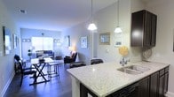 This thoughtfully designed single-floor residence showcases an open-concept layout that seamlessly connects the kitchen, dining, and living areas into one cohesive and functional space. The kitchen is a standout feature, appointed with sleek granite countertops, a stainless steel sink, and dark cabinetry that provides a striking contrast to the light stone surfaces. Elegant pendant lights hang above the kitchen island, which doubles as a breakfast bar with seating for multiple guests — ideal for casual dining or entertaining. Rich dark hardwood flooring flows throughout the main living areas, adding warmth and continuity to the space. The living room is generously proportioned, offering ample room for comfortable seating arrangements, and benefits from large windows that invite natural light to fill the interior, creating a bright and welcoming ambiance. Neutral gray walls provide a versatile backdrop that complements a range of furnishing styles. Decorative framed artwork adorns the walls, adding personality and a curated feel to the space. The overall atmosphere is modern yet inviting, with a clean aesthetic and well-considered design details that make this a comfortable and stylish place to call home.