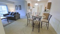 This single-floor residence features an open-concept floor plan that seamlessly connects the main living areas, creating a cohesive and functional living environment. The dining area serves as a central hub, accommodating a round glass-top table with elegant wrought-iron chairs and cushioned seats, illuminated by a pendant light fixture hanging overhead. The neutral carpeting throughout the living and dining spaces contributes to a warm, understated aesthetic.

Adjacent to the dining area, the living room offers a comfortable, intimate setting with a dark upholstered sofa and framed wall art, lending a relaxed atmosphere to the space. The open passageway connecting these areas enhances the sense of flow and accessibility throughout the home.

The kitchen is efficiently laid out with light wood cabinetry, white appliances including a range and refrigerator, and a countertop workspace well-suited for everyday cooking. A secondary kitchen counter area visible to one side provides additional prep and storage space. The overall color palette of soft blues, whites, and warm neutrals creates a calm, inviting ambiance across the entire residence.