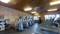 This fitness center is a thoughtfully designed, single-floor workout facility that combines functionality with a warm, welcoming ambiance. The space is defined by its striking wood-paneled ceiling, which lends a natural, lodge-like character to the room and sets it apart from typical gym environments. Recessed fluorescent lighting and a ceiling fan ensure the space remains bright and well-ventilated throughout use.

The main workout area is generously proportioned, offering ample room for a robust lineup of cardio equipment, including multiple elliptical trainers and a row of treadmills positioned along one side of the room. A dedicated strength and cable machine station is visible toward the rear of the space, providing options for resistance training alongside the cardio offerings.

Large mirrors line one wall, enhancing the sense of openness and allowing users to monitor their form during workouts. The flooring is a durable, dark-toned material well-suited for high-traffic fitness use. Natural light filters in through several windows, connecting the interior to the outdoors and contributing to an energizing atmosphere. The warm-toned walls complement the wood ceiling, creating a cohesive and inviting environment that encourages regular use. Overall, this fitness center is a well-rounded facility designed to support a variety of exercise routines.